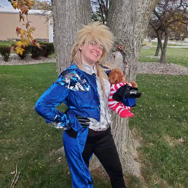 A person with spiky blonde hair, wearing a blue sequined jacket and black gloves, leans against a tree outdoors while…