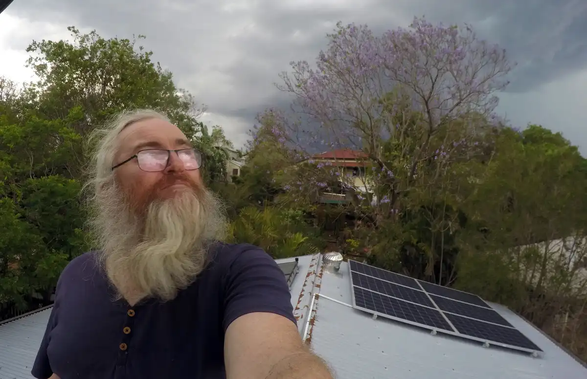 A man with a long white beard and glasses takes a selfie on a corrugated metal roof where solar panels are installed. Behind…