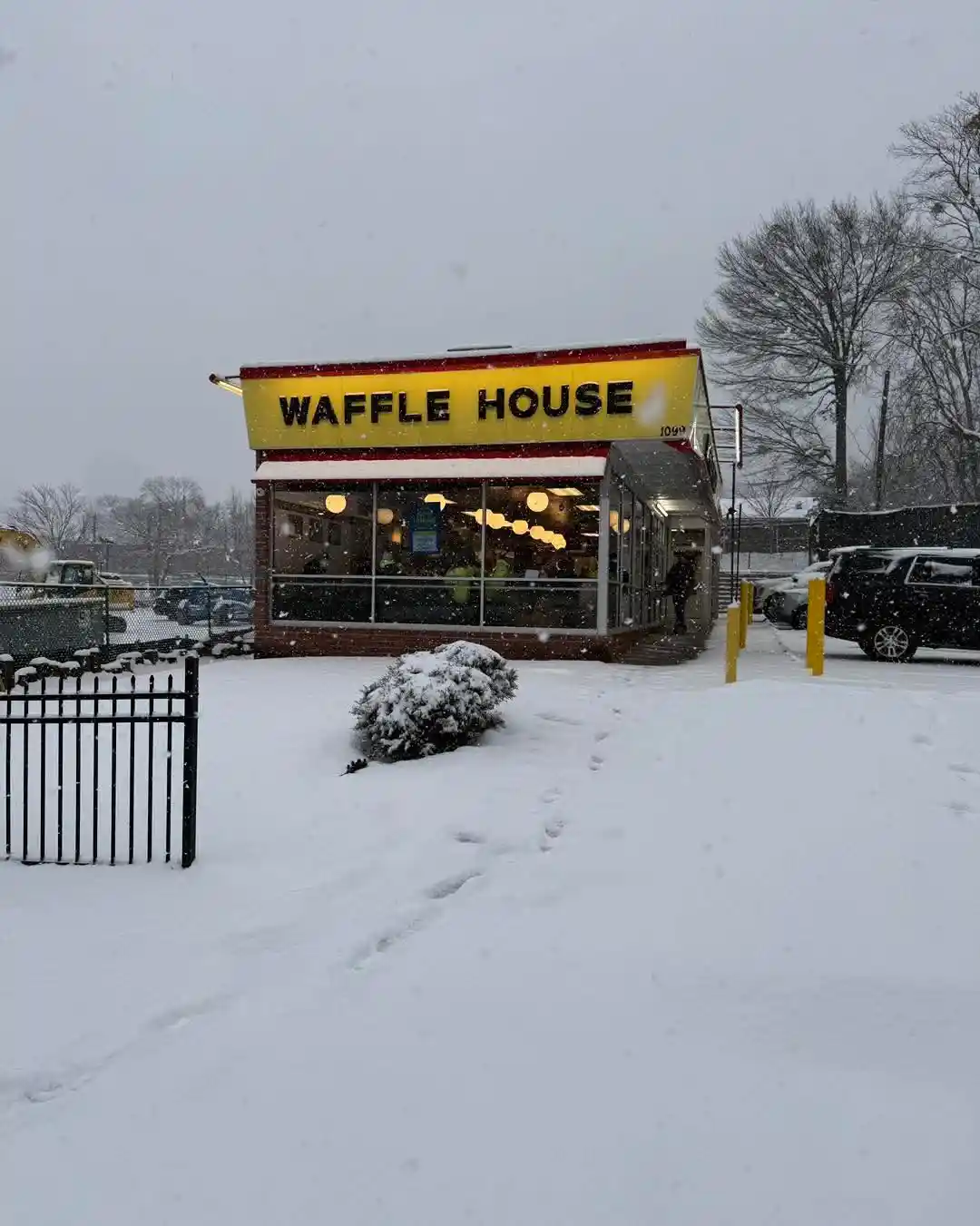 A snowy outdoor photo of a Waffle House restaurant with its iconic yellow sign visible above the entrance. The ground is…