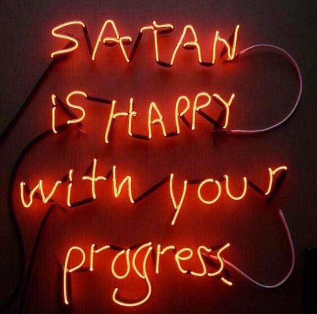 A red neon sign with glowing orange letters spells out 'SATAN IS HAPPY with your progress' in a handwritten style, mounted…