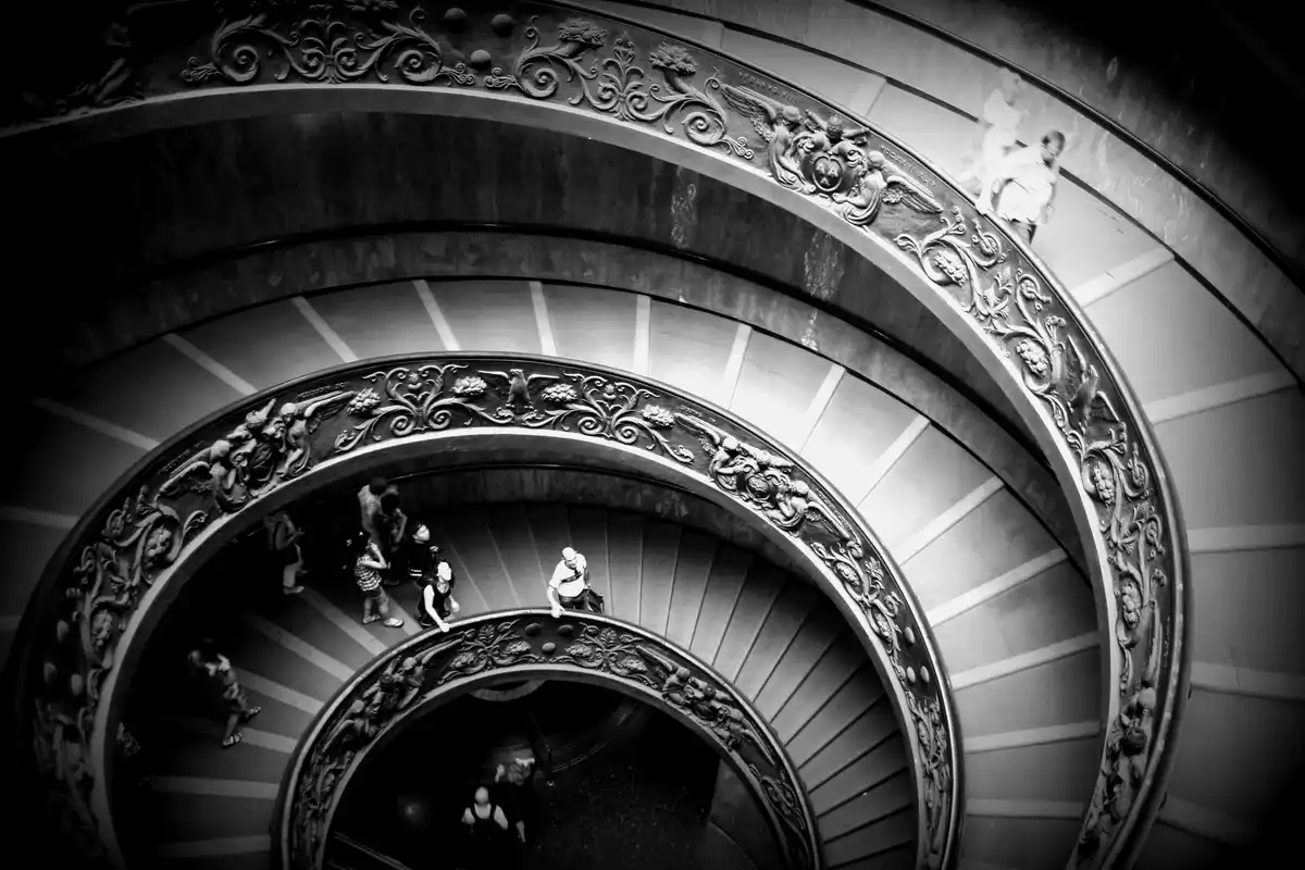 A black-and-white photo shows a grand, ornate spiral staircase with people walking on different levels. The text overlaid on…