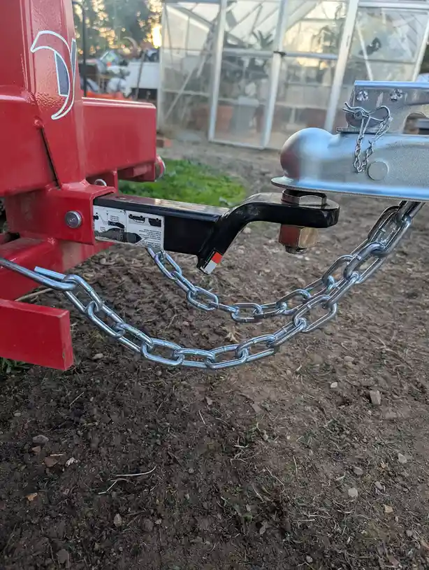 A close-up photo shows a red trailer hitch with a heavy-duty metal chain hanging loosely on the ground, attached to a silver…