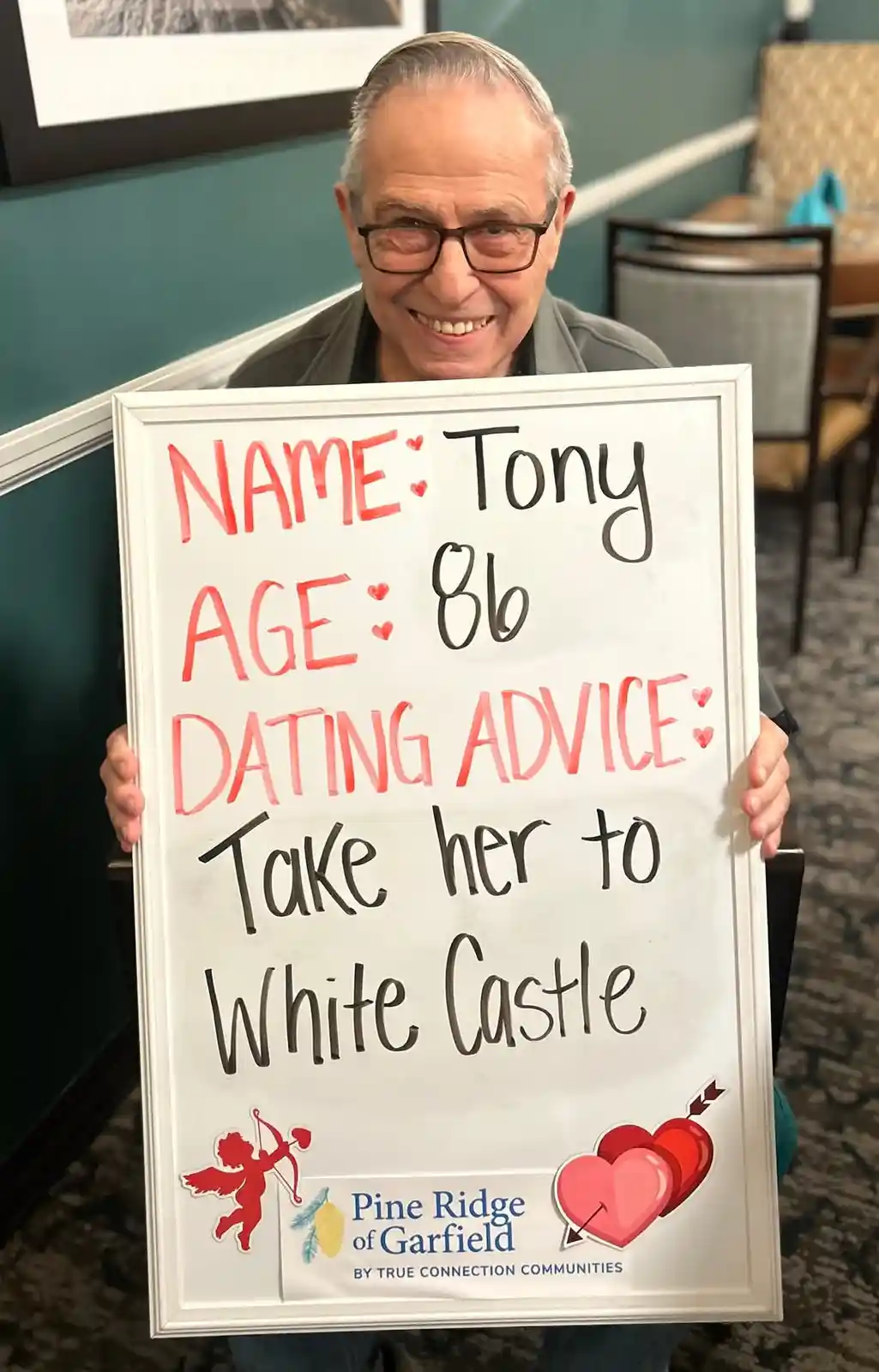 An elderly man with glasses smiles while holding a white-framed sign with handwritten text. The sign lists his name as Tony…