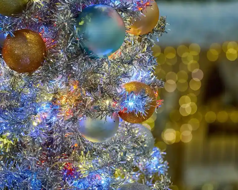 A close-up of a silver tinsel christmas tree decorated with shiny blue, gold, and white ornaments and twinkling lights, with…