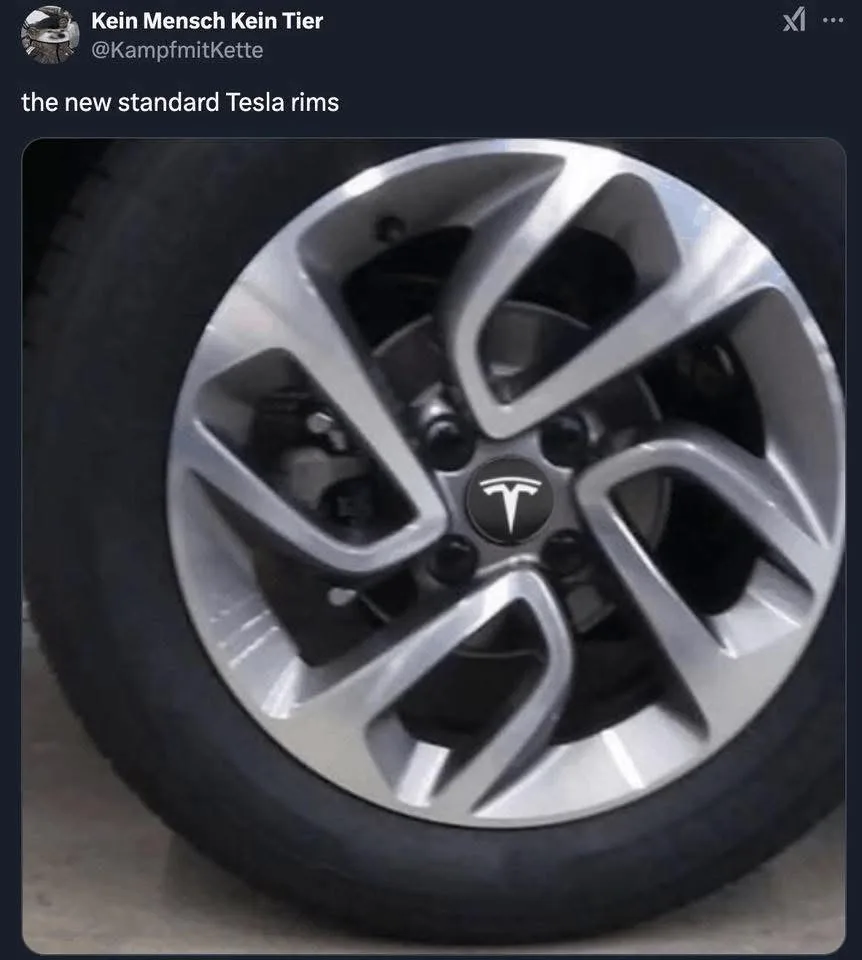 A close-up photo of a silver Tesla wheel rim with a black tire, showing the Tesla logo in the center. The overlaid text…