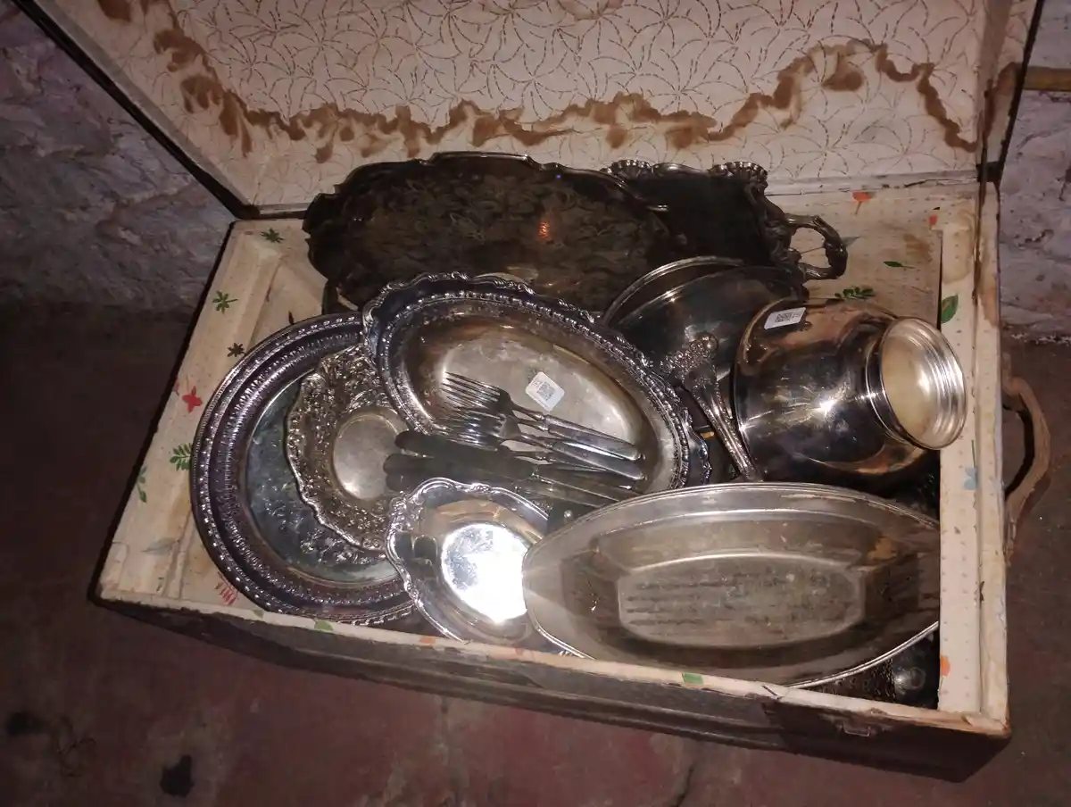 An open, worn wooden chest contains a jumble of tarnished silver plates, bowls, and forks, all piled together. The interior…