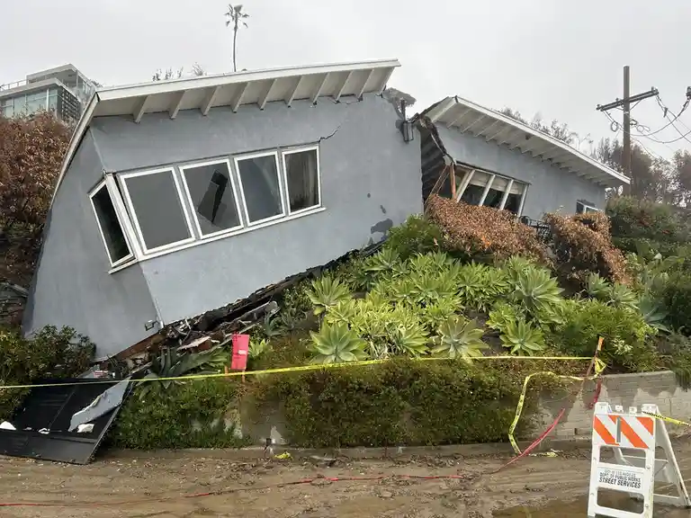 A light blue house is visibly tilted and damaged, sliding down a hillside with broken windows and exposed framing. Yellow…