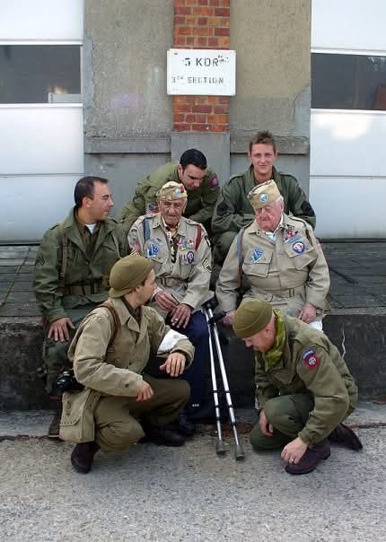 A group of seven men in military-style uniforms pose outdoors in front of a building with a sign reading '5 KDR 3rd…