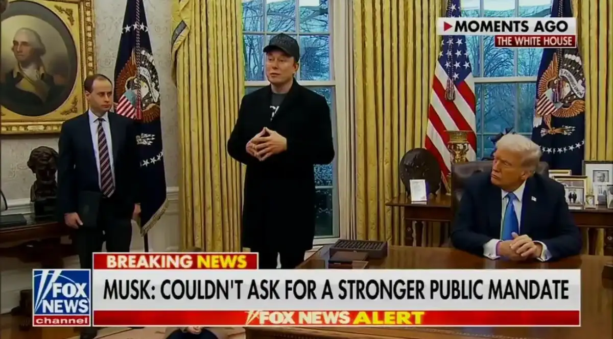 A still from a Fox News broadcast shows Elon Musk standing in the Oval Office, wearing a black jacket and cap, with Donald…