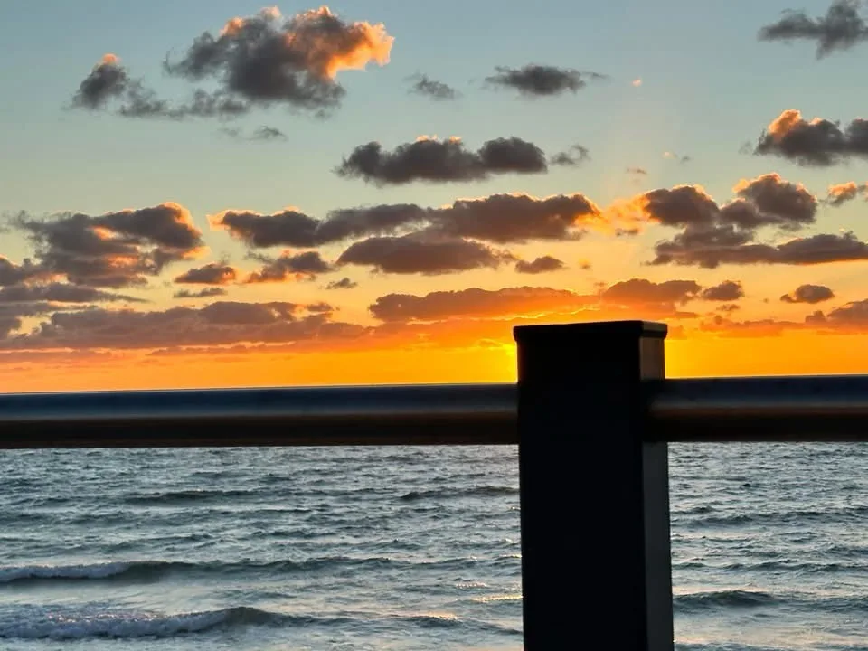 A serene sunset over the ocean with scattered clouds, viewed from behind a dark metal railing. The sky transitions from…