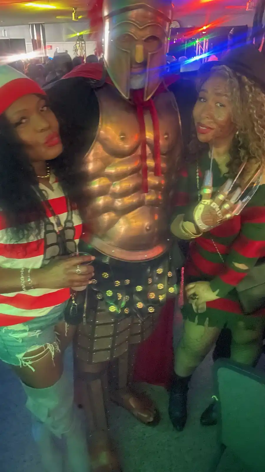 Three people pose together at a party: a person in a shiny gold gladiator costume with a helmet and red cape stands between…