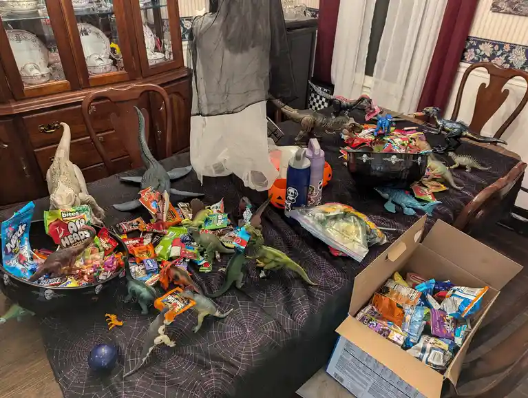 A dining table covered in a black spiderweb tablecloth is cluttered with numerous plastic dinosaur toys and bowls…