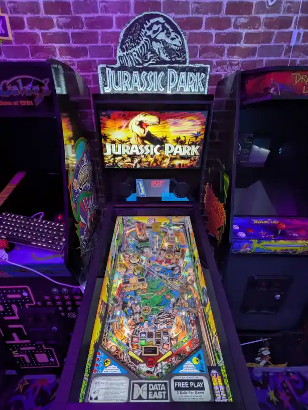 A brightly lit Jurassic Park pinball machine sits in an arcade, flanked by other games like Galaga and Dragon's Lair, with a…