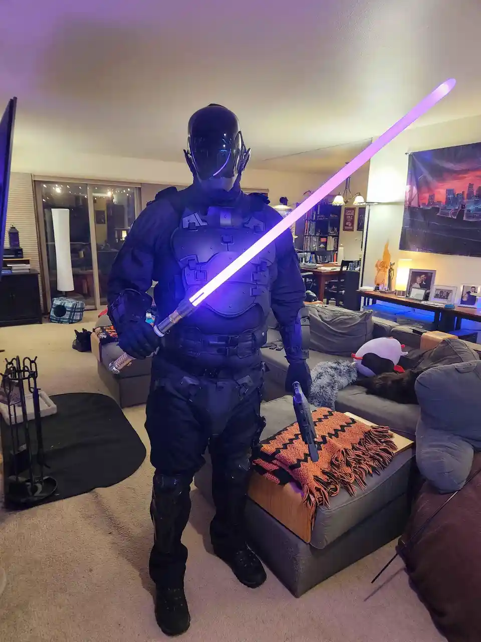 A person in a full-body black armored robot costume, complete with a helmet and glowing purple lightsaber, stands in a…