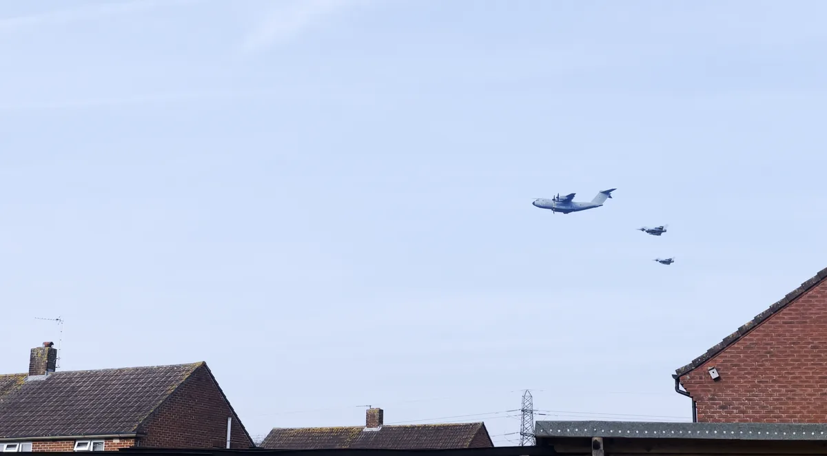 A photograph shows three military aircraft flying low over residential rooftops under a pale blue sky. A large grey…