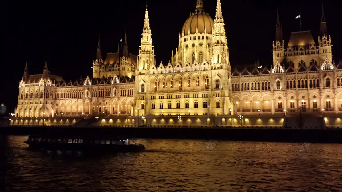 A nighttime photo of the illuminated Hungarian Parliament Building along the Danube River, with a boat visible in the…
