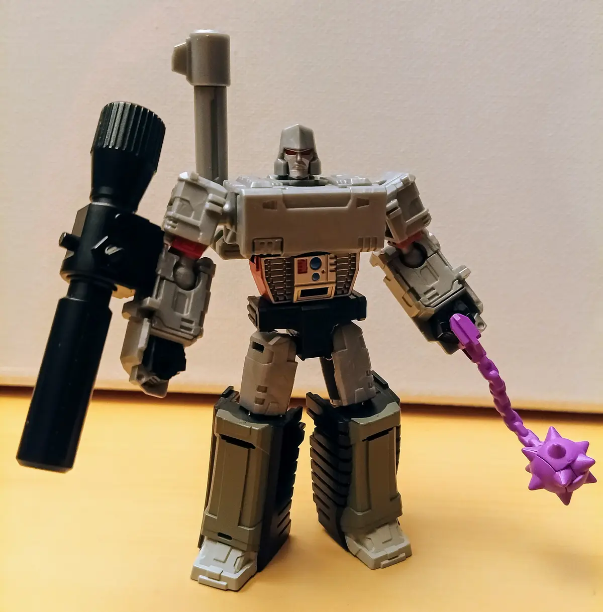 A gray and black transformer toy stands on a wooden surface against a beige wall, holding a large black weapon in one hand…