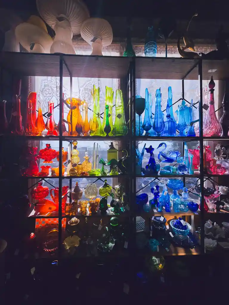 A dark room features shelves filled with colorful glassware, including vases, bottles, and decorative pieces in red, orange…