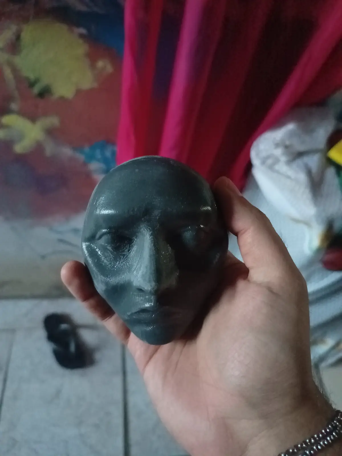 A hand holds a dark gray, 3D-printed or sculpted face with a blank, neutral expression. The face has visible layer lines…
