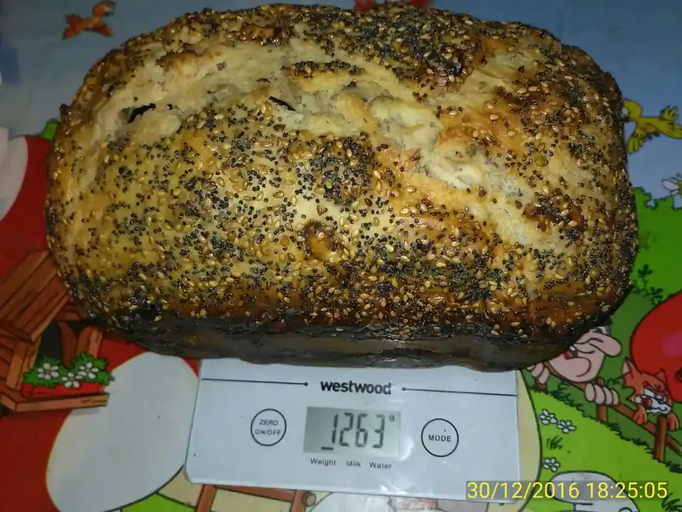 A large, golden-brown loaf of seeded bread sits on a digital kitchen scale displaying '1263g'. The bread is topped with a…