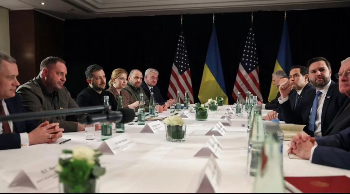 A formal meeting of international leaders seated at a long table with American and Ukrainian flags behind them. The overlaid…