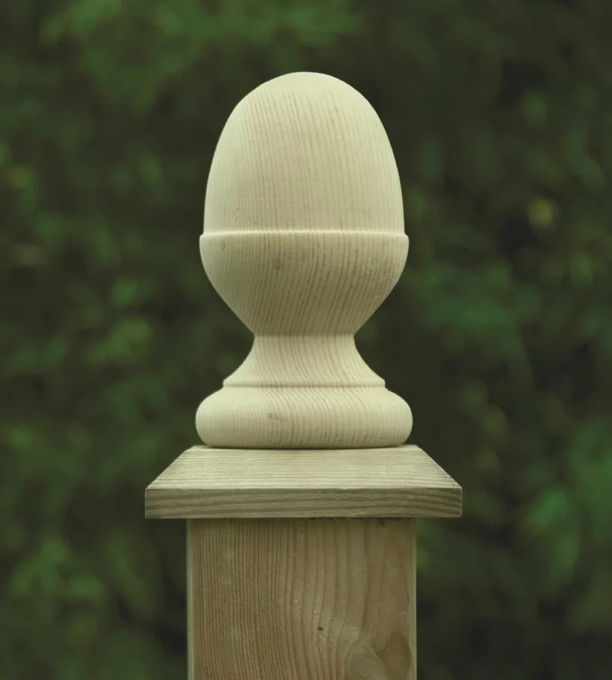 A close-up of a light-colored wooden fence post with a carved, egg-shaped finial on top, set against a blurred green foliage…