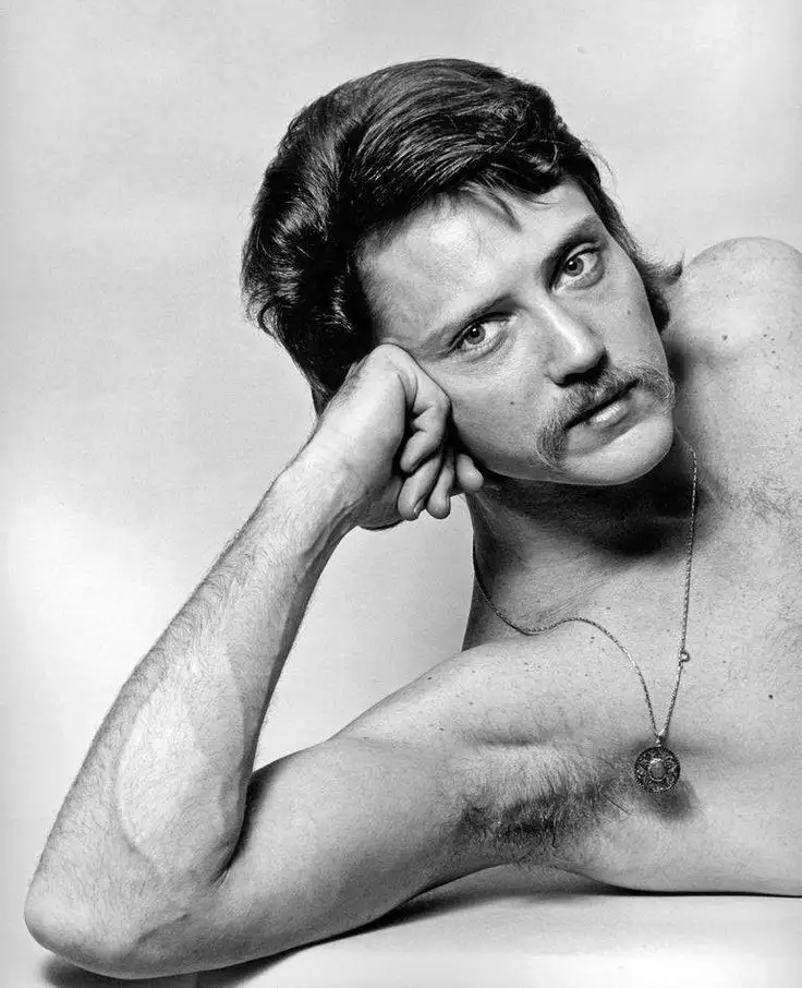 A black-and-white photo of a shirtless man with a mustache and styled hair, resting his head on his hand while looking…