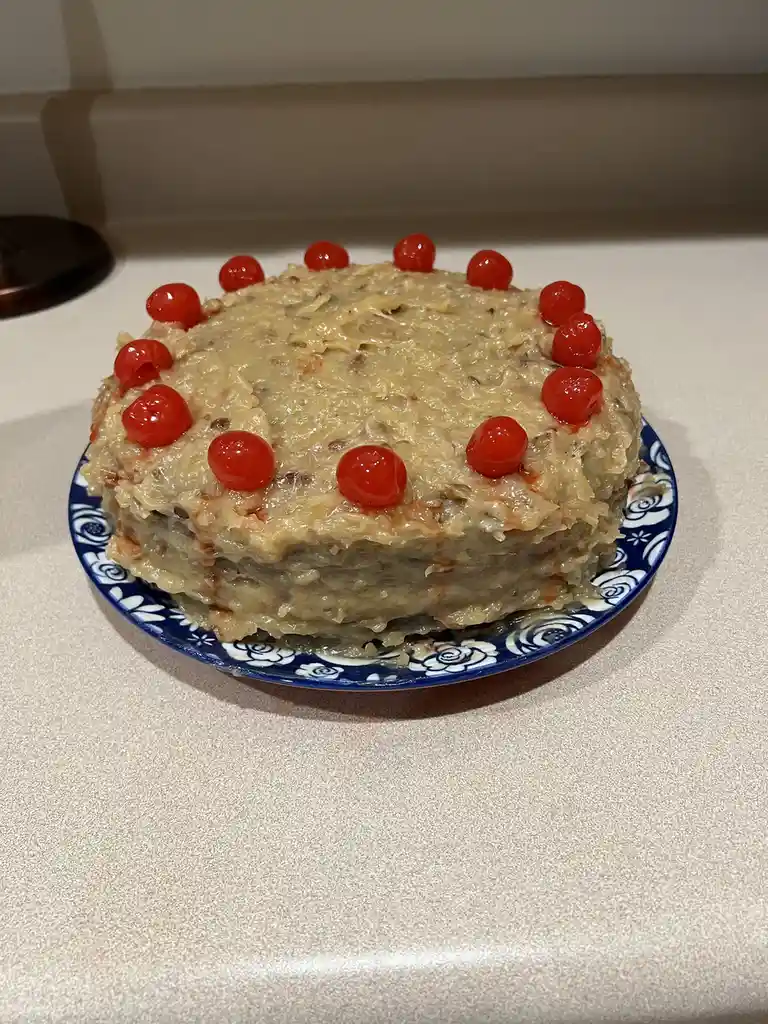 A round, multi-layered dessert with a thick, textured, light-brown frosting and bright red maraschino cherries arranged in a…