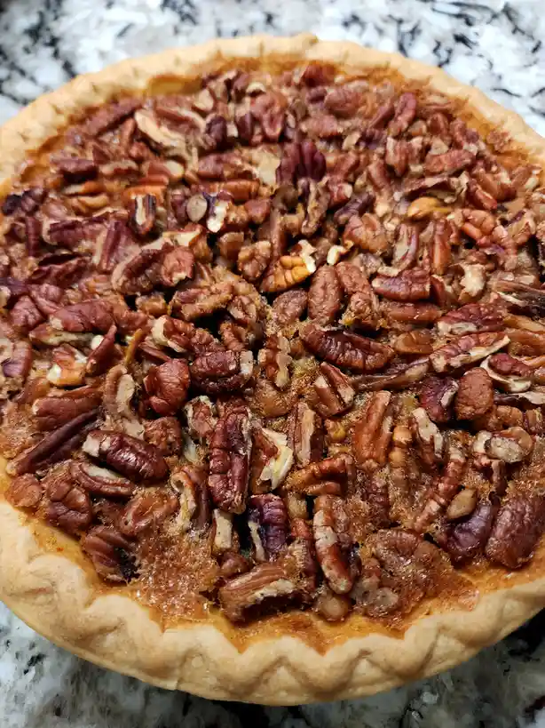 A close-up photo of a freshly baked pecan pie with a golden-brown crust and a top generously covered with toasted pecan…