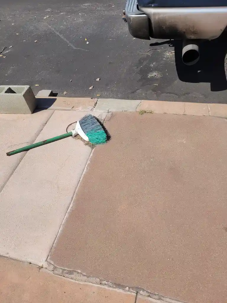 A broom with a green handle and two-tone bristles (gray and teal) lies on a sidewalk next to a curb, with a car's exhaust…
