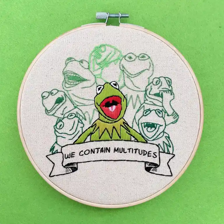 An embroidery hoop displays a stitched image of Kermit the Frog in the center, surrounded by outlines of other Muppet…
