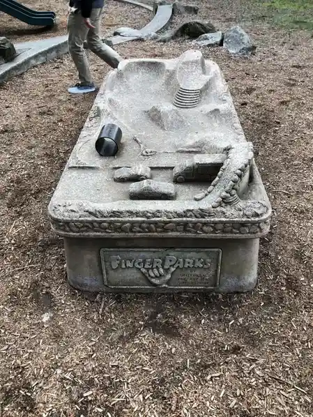 A concrete sandbox at a playground features molded car and truck shapes, a spiral ramp, and a black pipe, with a person…