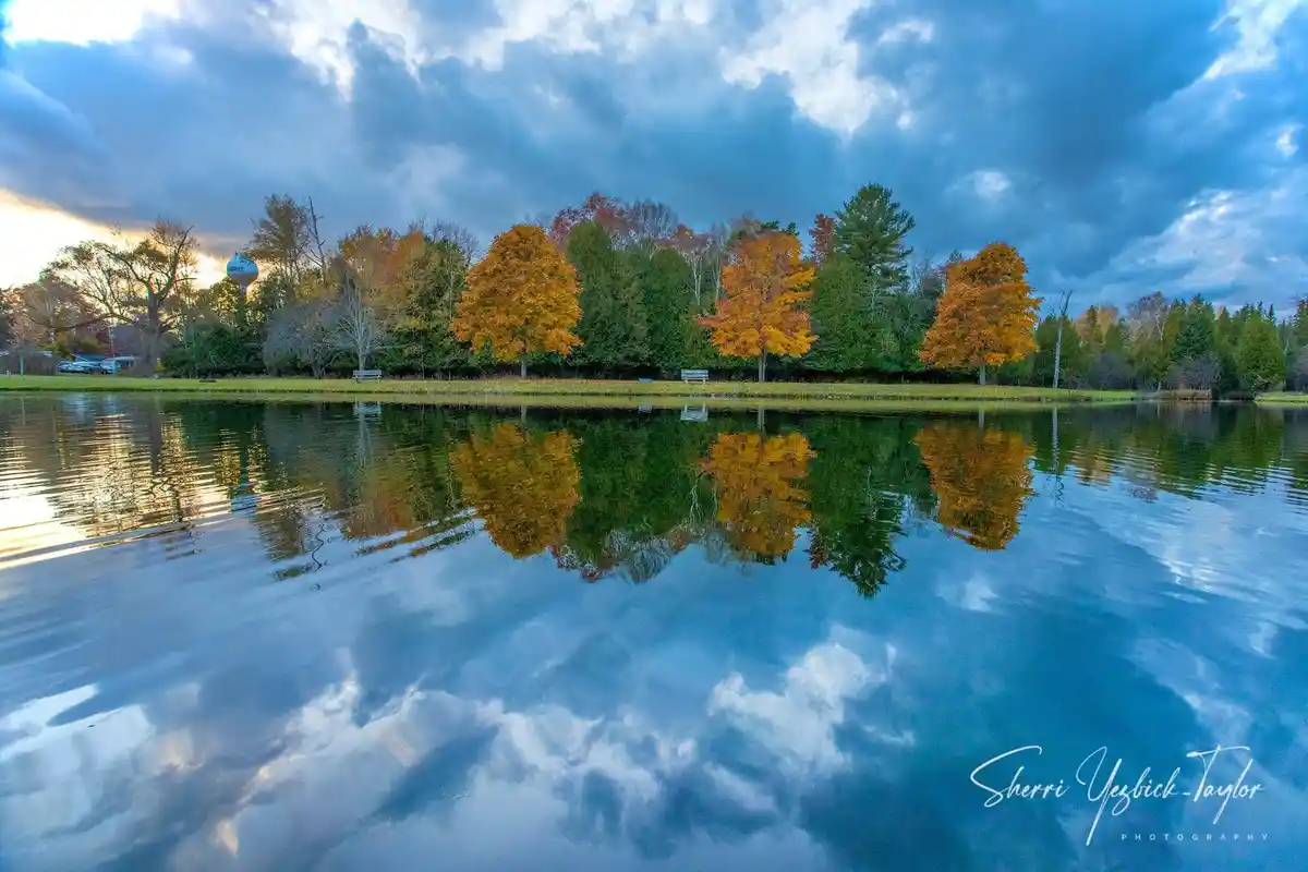 A serene autumn landscape shows a calm lake reflecting vibrant orange and green trees under a dramatic cloudy sky. The image…