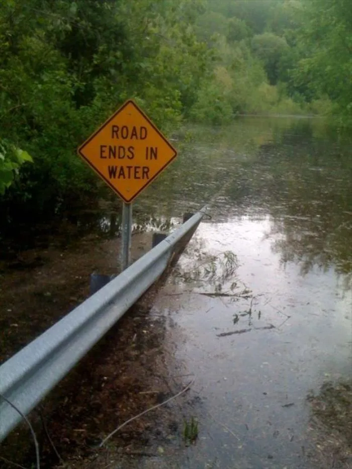 A yellow diamond-shaped road sign reading 'ROAD ENDS IN WATER' stands beside a flooded road, with water visibly covering the…