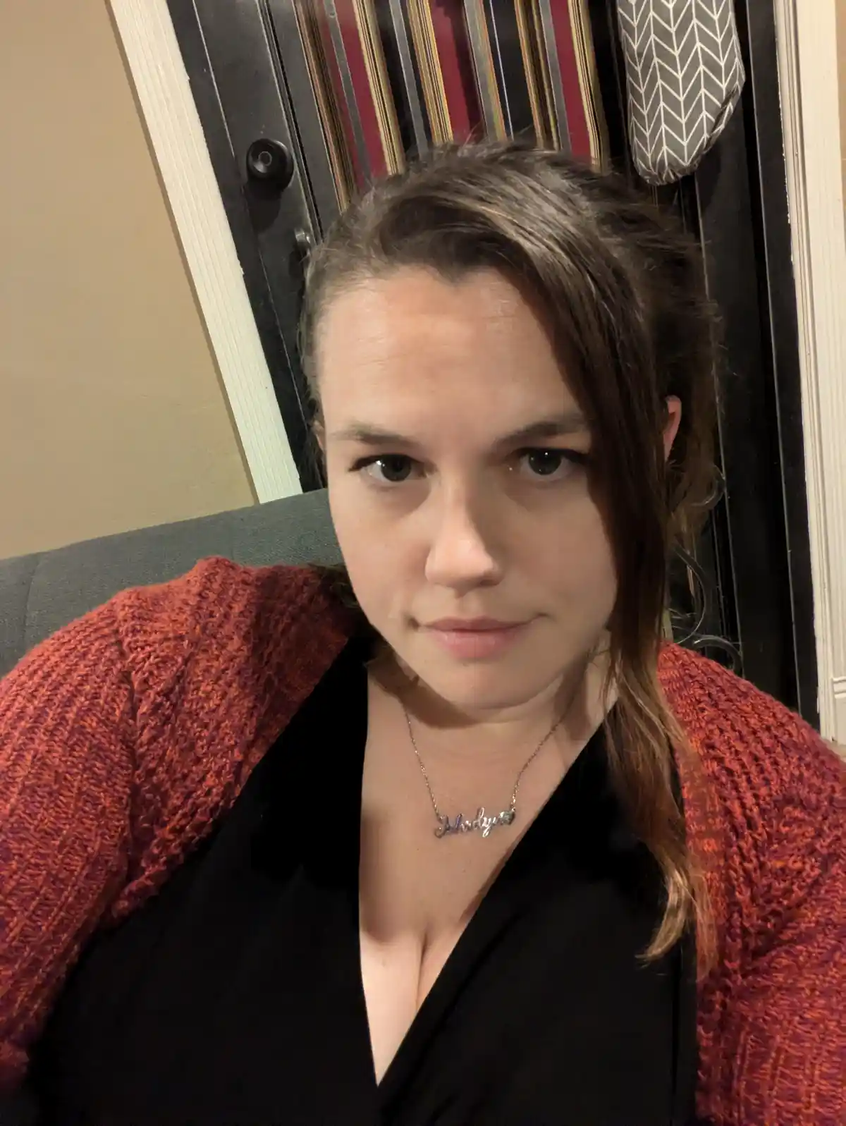 A woman with brown hair wearing a red knitted cardigan over a black top sits on a gray couch, looking directly at the camera…