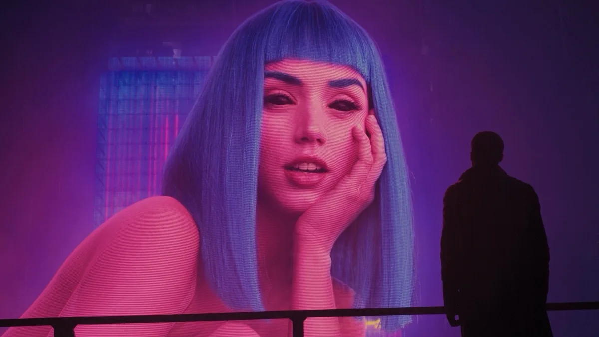 A person's silhouette stands before a large, glowing digital billboard displaying a woman with blue hair and a hand on her…