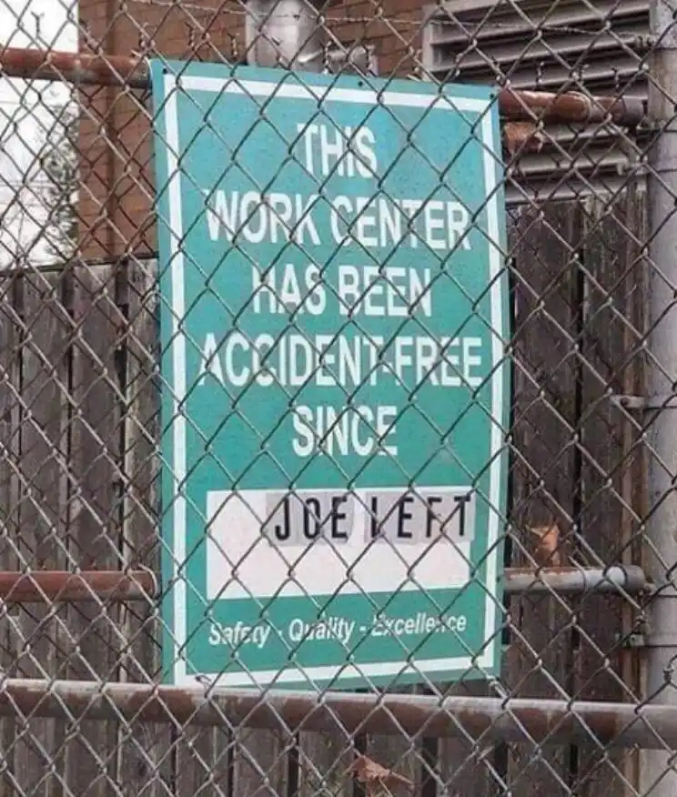 A green safety sign behind a chain-link fence reads 'THIS WORK CENTER HAS BEEN ACCIDENT-FREE SINCE' with the word 'JOE LEFT'…
