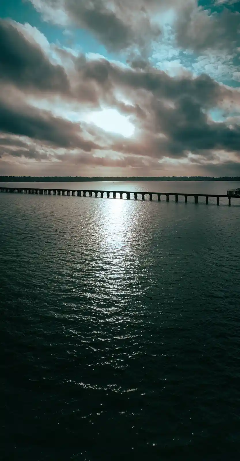 An aerial view of a long pier extending into a dark body of water under a dramatic, cloudy sky with sunlight breaking…