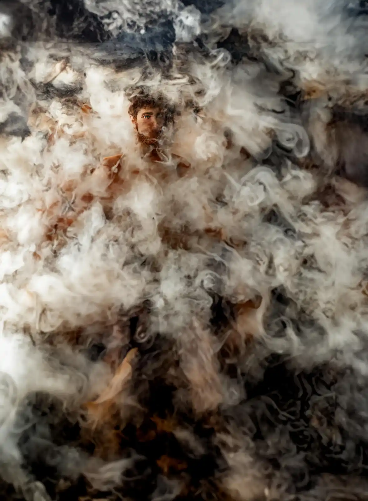 A shirtless man with dark hair and a beard is partially obscured by swirling white and gray smoke, with only his face and…