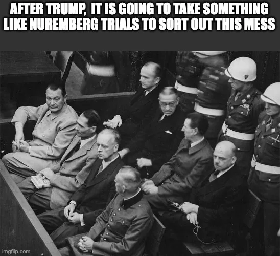 A black and white historical photograph shows several men in military-style uniforms seated in a courtroom, flanked by…