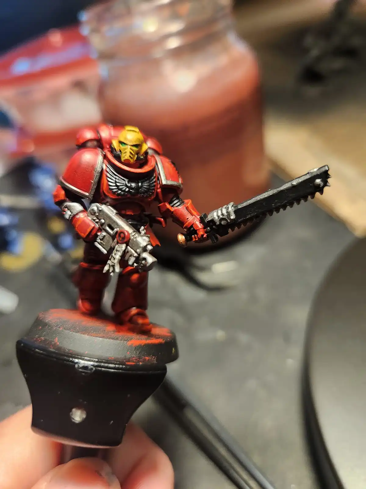 A hand holds a painted miniature of a red-armored Space Marine with a yellow helmet and green eyes, wielding a chainsword…