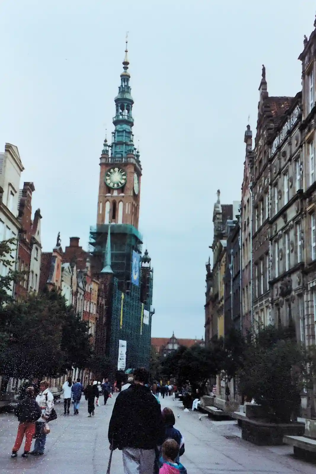 A street view looking down a European-style pedestrian street toward a tall clock tower partially covered in green…