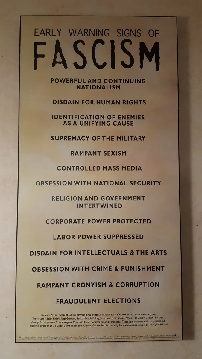 A printed poster titled 'EARLY WARNING SIGNS OF FASCISM' lists characteristics like 'POWERFUL AND CONTINUING NATIONALISM'…