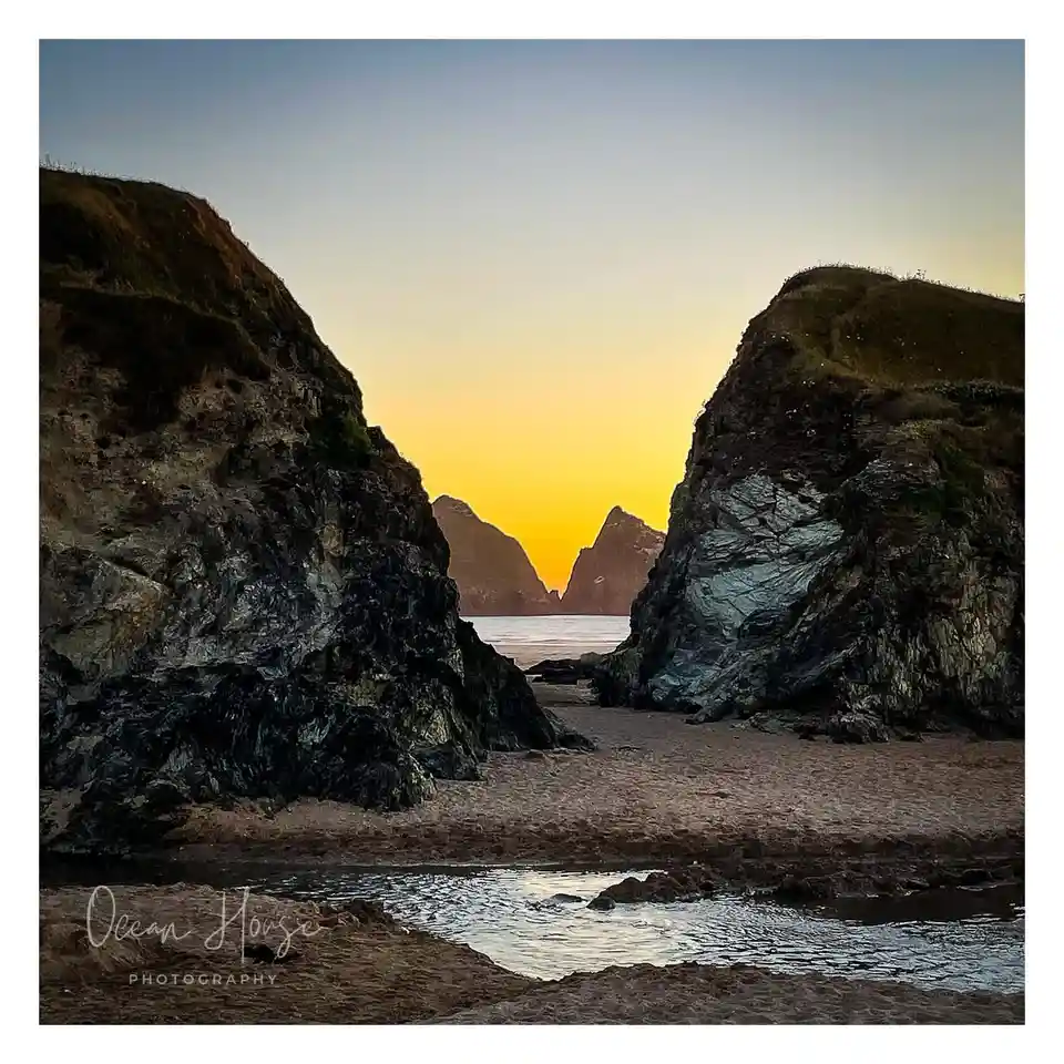 A serene landscape photo shows a narrow beach flanked by large, dark, mossy cliffs, with a golden sunset visible through a…