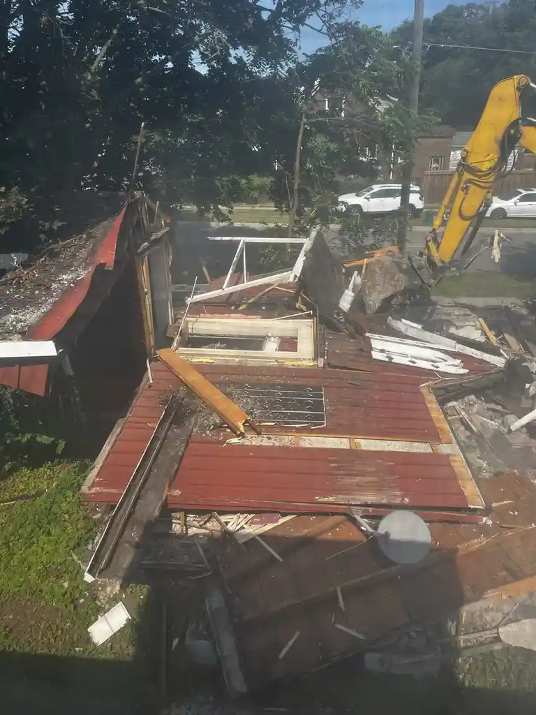 A partially demolished house with red metal roofing and scattered debris is shown from an elevated angle, with a yellow…