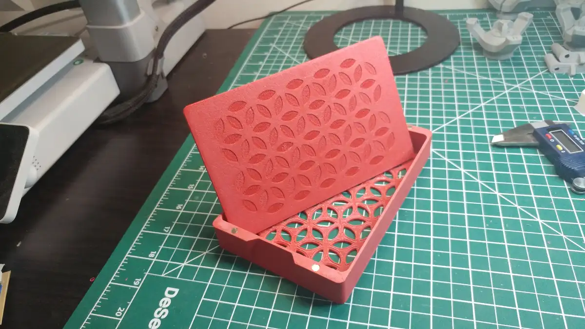 A red 3D-printed box with a lid featuring a repeating leaf-like pattern sits open on a green cutting mat. The interior has a…