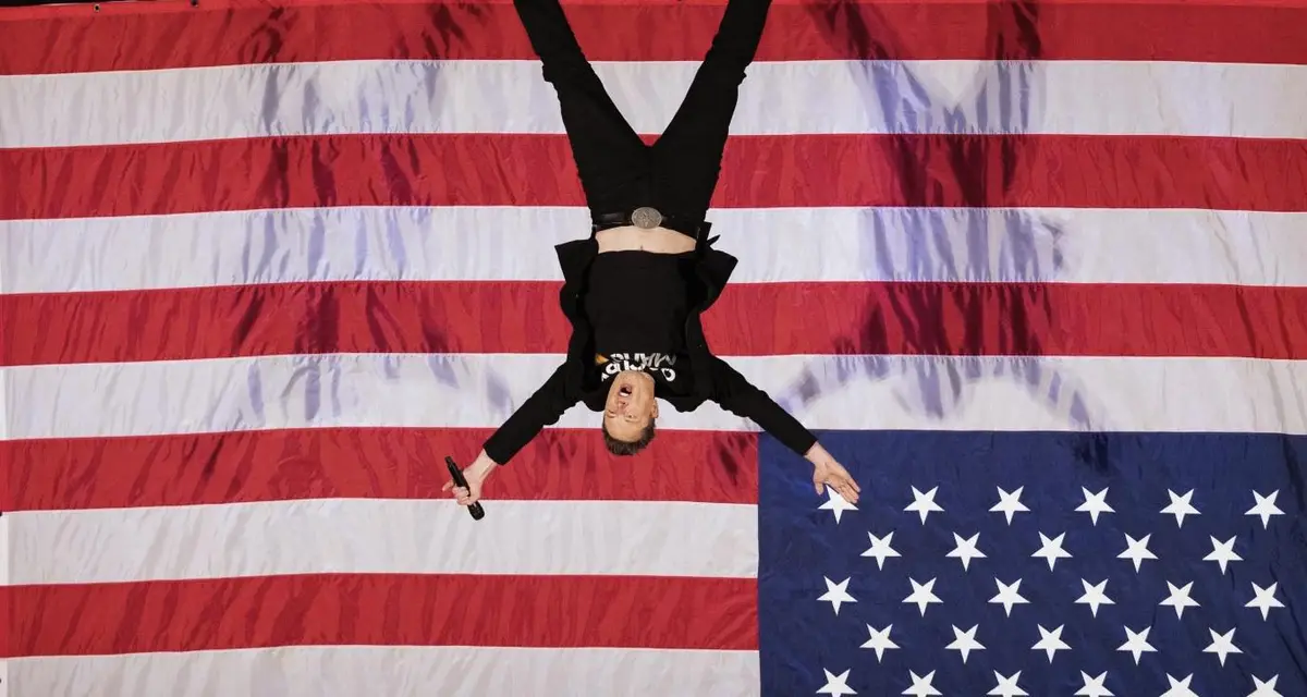 a person wearing a black shirt and pants is lying upside down on a large american flag, holding a microphone in one hand…