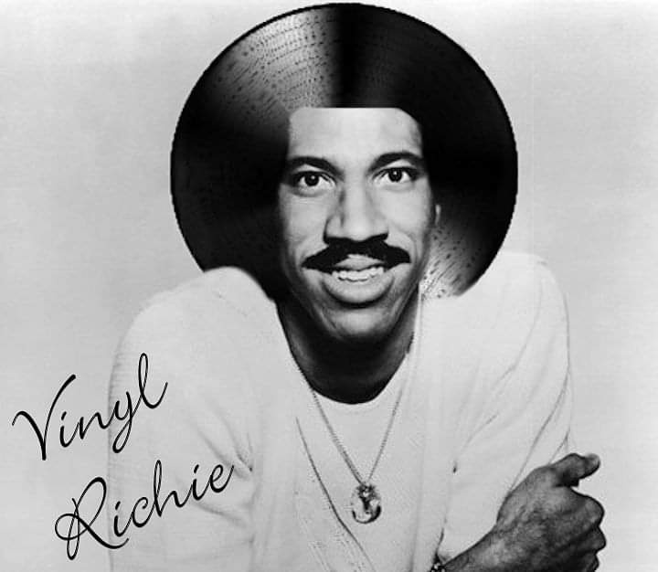 A black and white photo of a smiling man with a mustache, wearing a light-colored shirt and a necklace. His head is…
