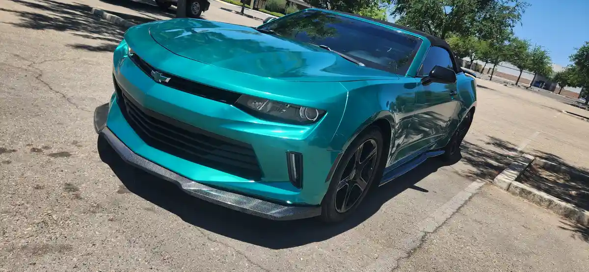 A shiny teal Chevrolet Camaro convertible is parked in a sunny lot, its glossy paint reflecting trees and sky. The car has…