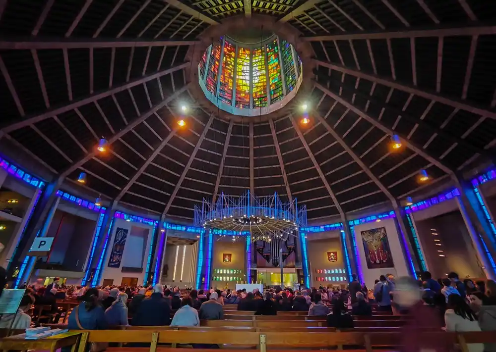 a wide-angle photo of a large, modern church interior filled with congregants seated in pews, looking toward the altar. The…